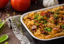 A freshly baked chicken orzo dish in a black casserole dish, garnished with herbs and surrounded by fresh tomatoes, garlic, and peas on a rustic wooden table.