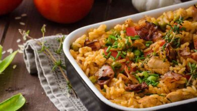 A freshly baked chicken orzo dish in a black casserole dish, garnished with herbs and surrounded by fresh tomatoes, garlic, and peas on a rustic wooden table.