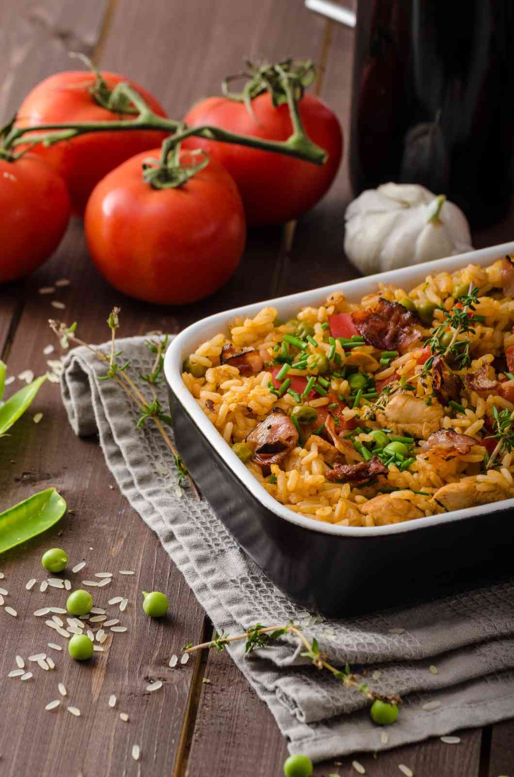 A freshly baked chicken orzo dish in a black casserole dish, garnished with herbs and surrounded by fresh tomatoes, garlic, and peas on a rustic wooden table.