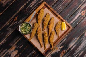 A rustic wooden cutting board displaying golden-brown, crispy fried smelt fish, arranged neatly on parchment paper. A small metal bowl of herb-infused butter sauce sits to the side, alongside a fresh lemon wedge for garnish. The dark wooden table background enhances the rich textures and colors of the dish.