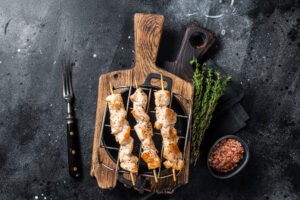 Three grilled chicken skewers with a golden-brown, slightly charred exterior are placed on a small round grill over a rustic wooden cutting board. Fresh sprigs of thyme, a small bowl of pink Himalayan salt, and a vintage fork with a black handle are arranged nearby on a dark textured background.