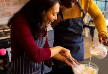 A cheerful couple wearing aprons is baking together in a warm, rustic kitchen. The woman is whisking a mixture in a glass bowl while the man pours milk. Fresh eggs, flour, and baking tools are arranged on the black countertop, creating a cozy and inviting cooking atmosphere