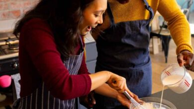 A cheerful couple wearing aprons is baking together in a warm, rustic kitchen. The woman is whisking a mixture in a glass bowl while the man pours milk. Fresh eggs, flour, and baking tools are arranged on the black countertop, creating a cozy and inviting cooking atmosphere