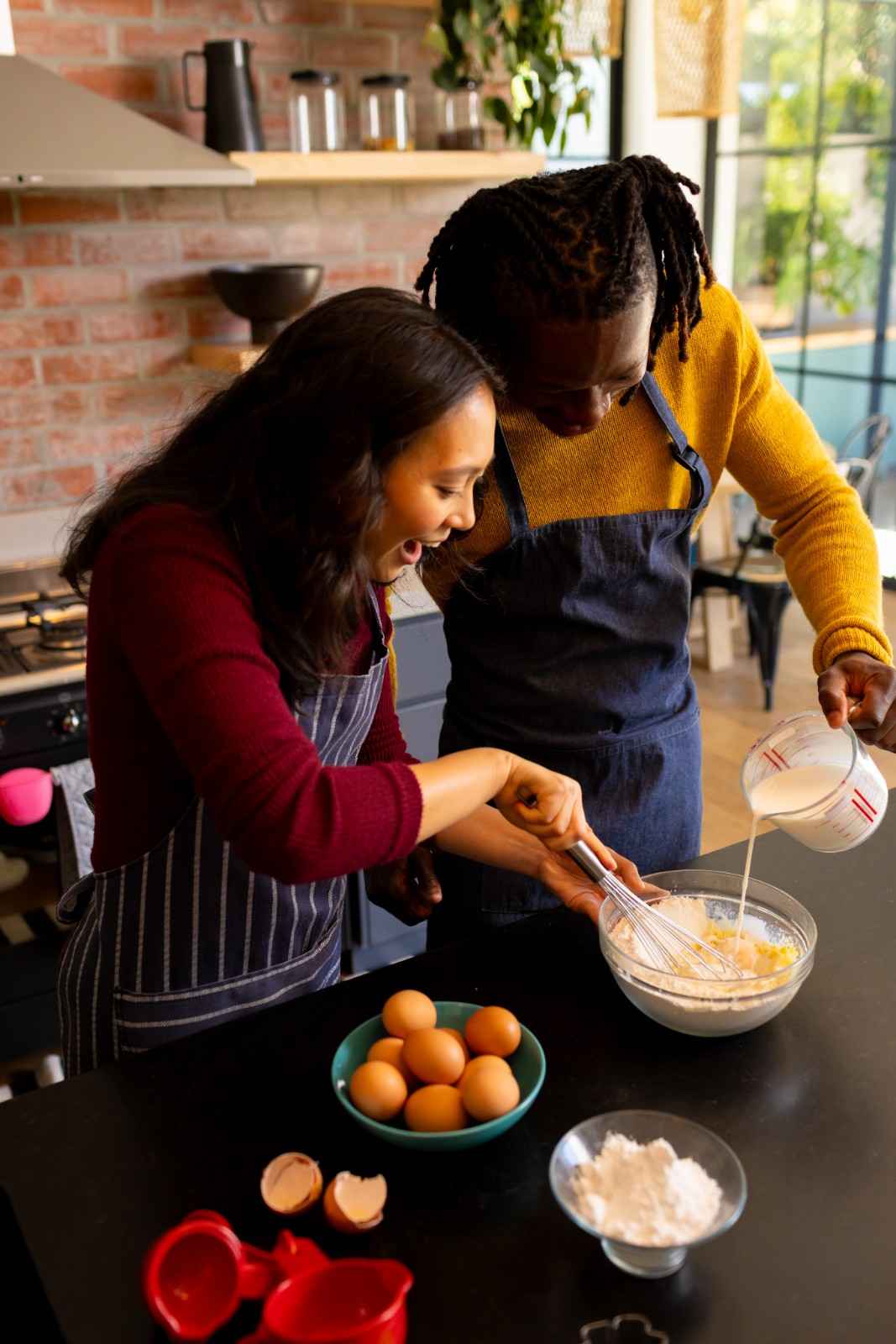 A cheerful couple wearing aprons is baking together in a warm, rustic kitchen. The woman is whisking a mixture in a glass bowl while the man pours milk. Fresh eggs, flour, and baking tools are arranged on the black countertop, creating a cozy and inviting cooking atmosphere