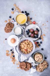 A beautifully arranged breakfast spread featuring a bowl of crunchy granola with dried fruit, a cup of milk, an egg, a plate of chocolate truffles with blueberries, chocolate chip cookies, and a warm beverage. The rustic setup on a textured gray background is complemented by scattered granola crumbs and fresh berries, creating a cozy and inviting morning scene.