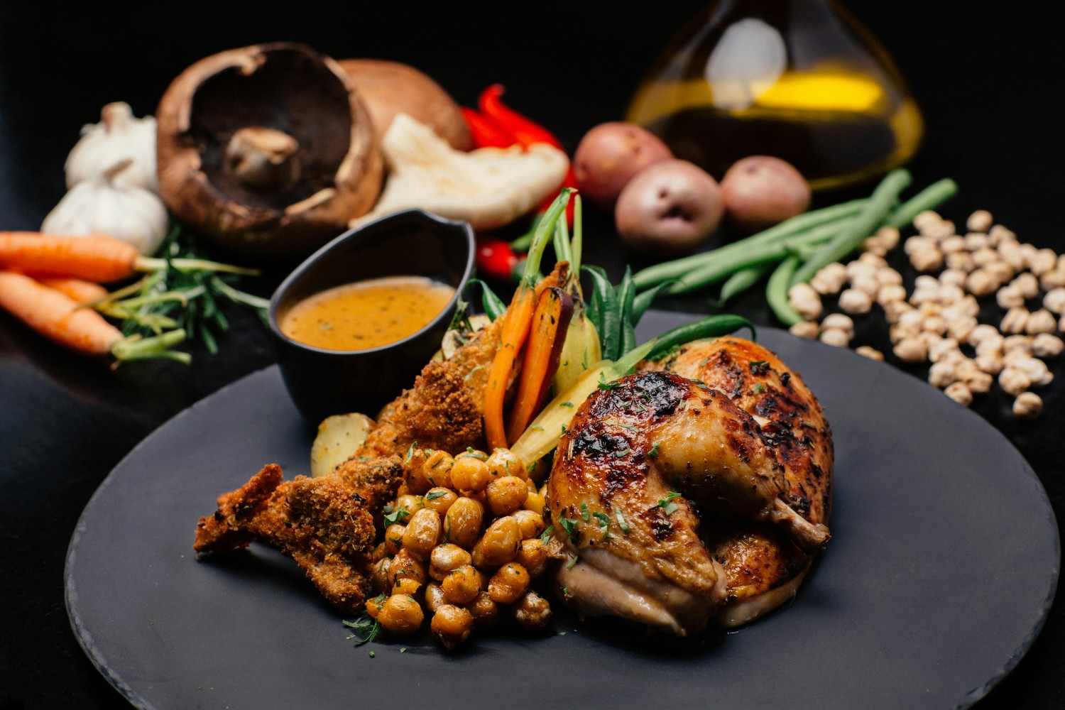 A beautifully plated garlic parmesan chicken dish served with crispy roasted chickpeas, breaded chicken, grilled vegetables, and a side of flavorful dipping sauce. Fresh ingredients like garlic, mushrooms, carrots, green beans, potatoes, and chickpeas are arranged in the background.