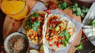 A close-up of two freshly made chicken tacos served on a wooden platter. The tacos feature soft flour tortillas filled with shredded chicken, cabbage, corn, salsa, guacamole, and pomegranate seeds, topped with fresh cilantro. Around the platter, there are lime wedges, a bowl of seasoning, a glass of citrus drink with a chili rim, and scattered herbs and spices, creating a vibrant and flavorful food presentation.