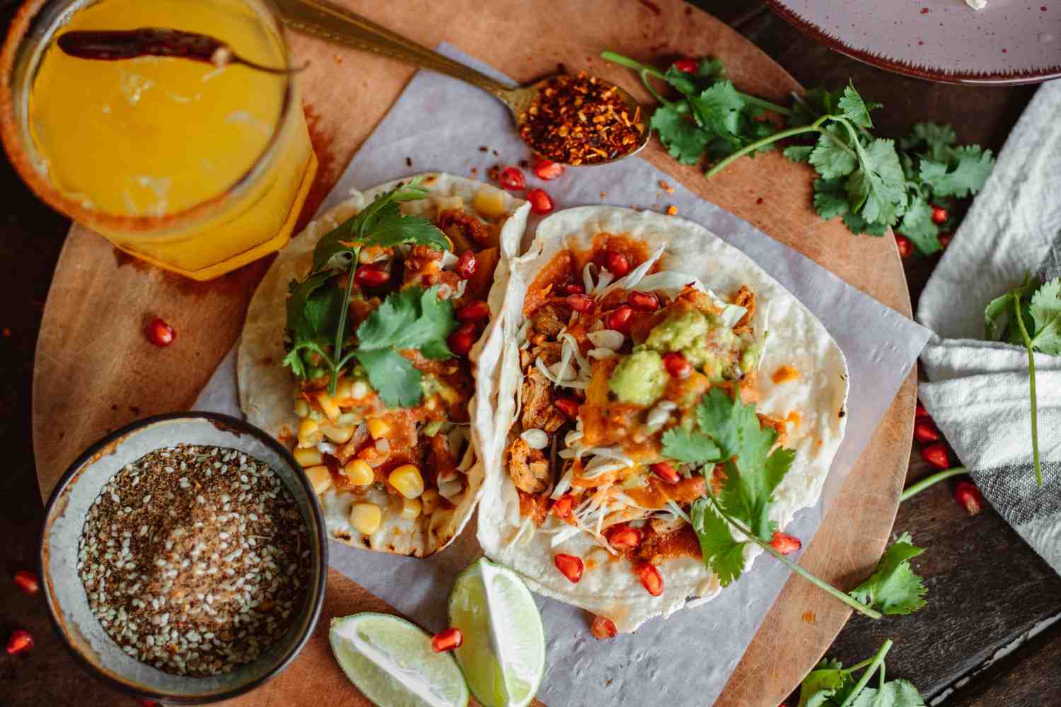 A close-up of two freshly made chicken tacos served on a wooden platter. The tacos feature soft flour tortillas filled with shredded chicken, cabbage, corn, salsa, guacamole, and pomegranate seeds, topped with fresh cilantro. Around the platter, there are lime wedges, a bowl of seasoning, a glass of citrus drink with a chili rim, and scattered herbs and spices, creating a vibrant and flavorful food presentation.