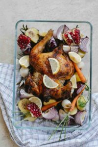 A golden-brown roasted chicken in a glass baking dish, garnished with lemon slices and surrounded by fresh vegetables, including carrots, red onions, garlic, and potatoes. The dish also contains pomegranate and roasted pear for a flavorful twist. Placed on a checkered kitchen towel for a rustic presentation.