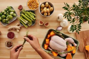 A top-down view of fresh ingredients for a roast chicken meal, including a raw whole chicken in a baking dish with rosemary and butternut squash. Surrounding the dish are bowls of potatoes, chickpeas, brussels sprouts, celery, spices, garlic, ginger, and figs, all neatly arranged on a wooden kitchen countertop. A person's hands are preparing rosemary for seasoning.