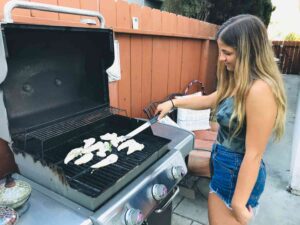 A young woman with long blonde hair, wearing a sleeveless top and denim shorts, is grilling chicken on a gas barbecue in an outdoor backyard setting. She is using tongs to turn the chicken pieces, which are seasoned with herbs. The grill is open, revealing the food cooking on the grates. A wooden fence, patio furniture, and a cushion with text are visible in the background.